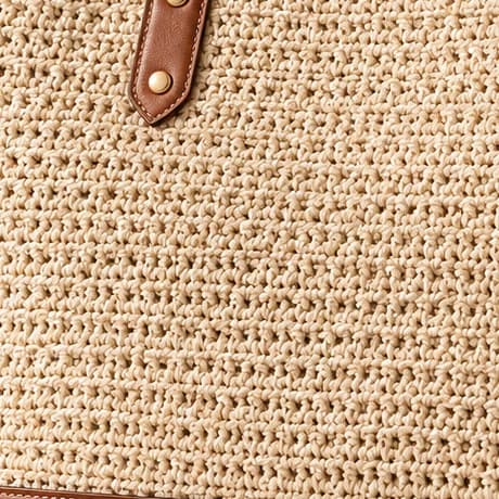 Close-up of hand-worked cotton crochet, stitch by stitch, with a cognac leather tab and a warm gold rivet